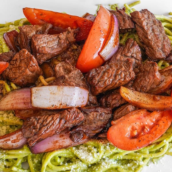 Peruvian Lomo Saltado with steak, tomatoes, and onions over green spaghetti, garnished with cheese.