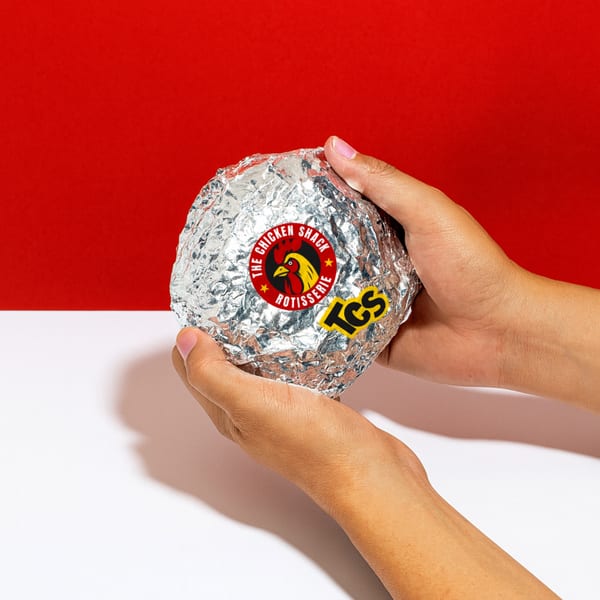 Hands holding a foil-wrapped Peruvian chicken from The Chicken Shack against a red and white background.