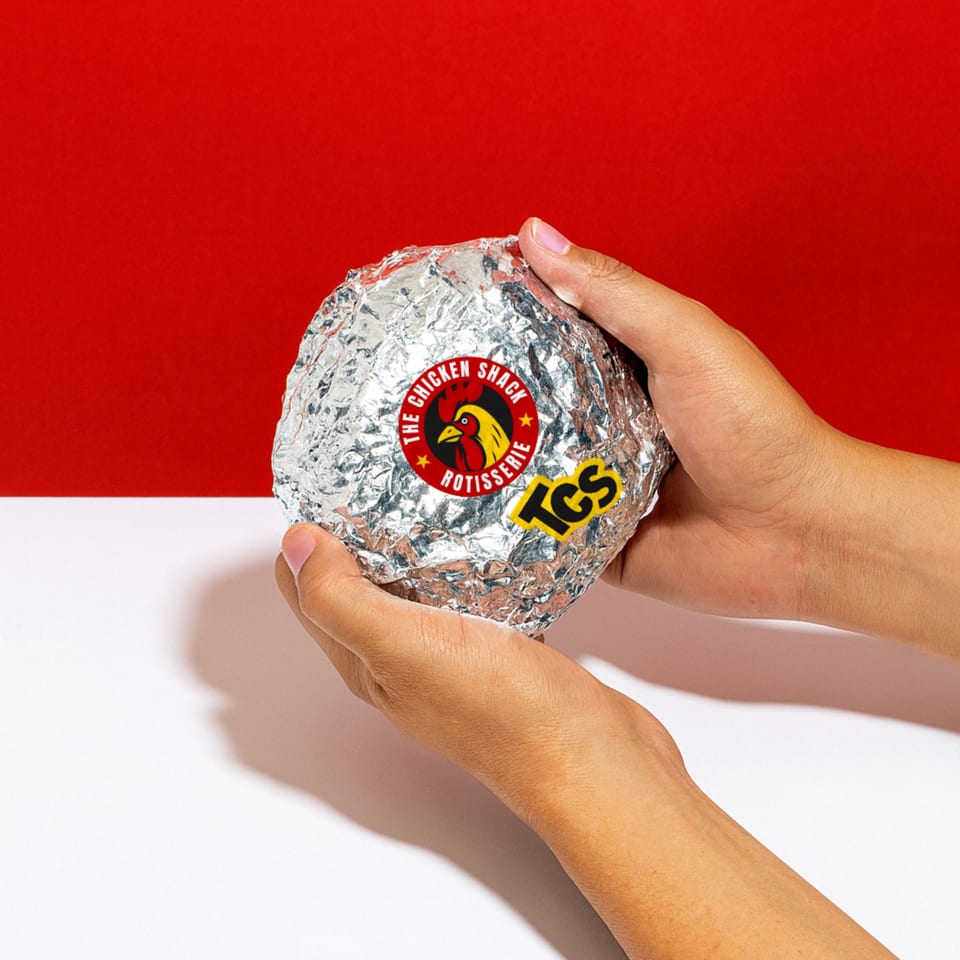 Hands holding a foil-wrapped Peruvian chicken from The Chicken Shack against a red and white background.