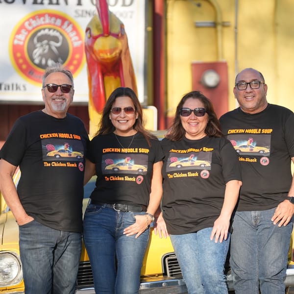Four smiling people in Chicken Shack t-shirts stand in front of a yellow car and chicken statue.