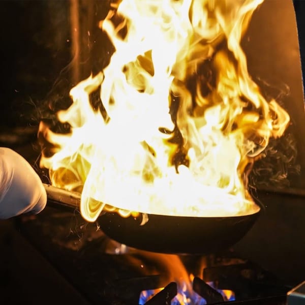 A chef's gloved hand holds a pan engulfed in bright yellow and orange flames over a stove.